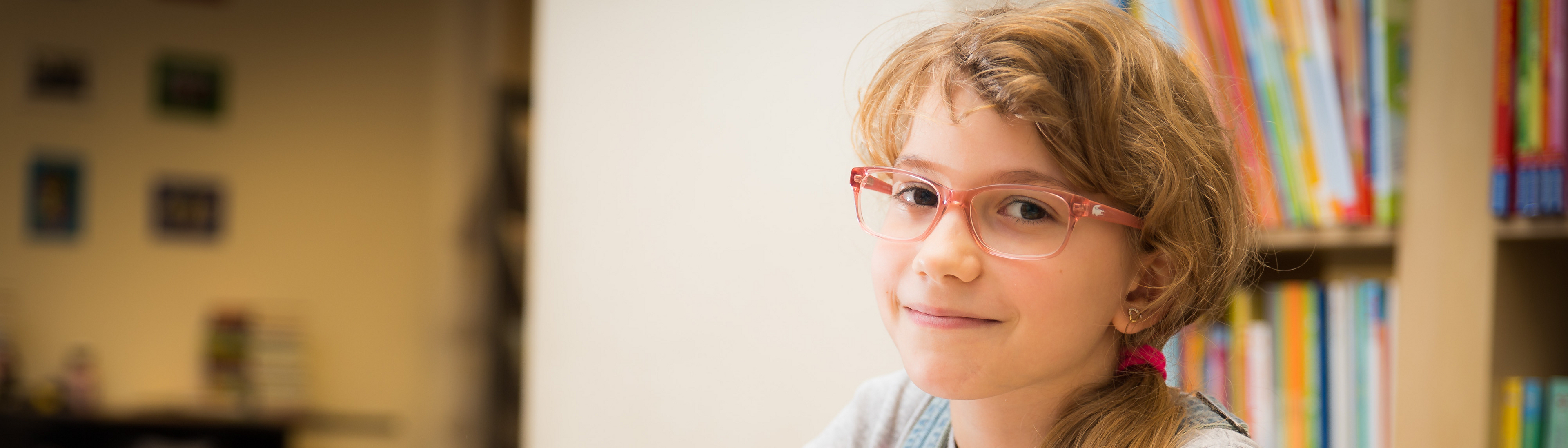 Mädchen mit roter Brille und Zopf, helle Haare, sitzt an Schreibtisch, davor Hefte, hält Stift in Hand und blickt seitlich in Kamera, im Hintergrund Bücherregal mit Büchern 