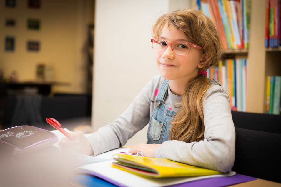 Mädchen mit roter Brille und Zopf, helle Haare, sitzt an Schreibtisch, davor Hefte, hält Stift in Hand und blickt seitlich in Kamera, im Hintergrund Bücherregal mit Büchern 