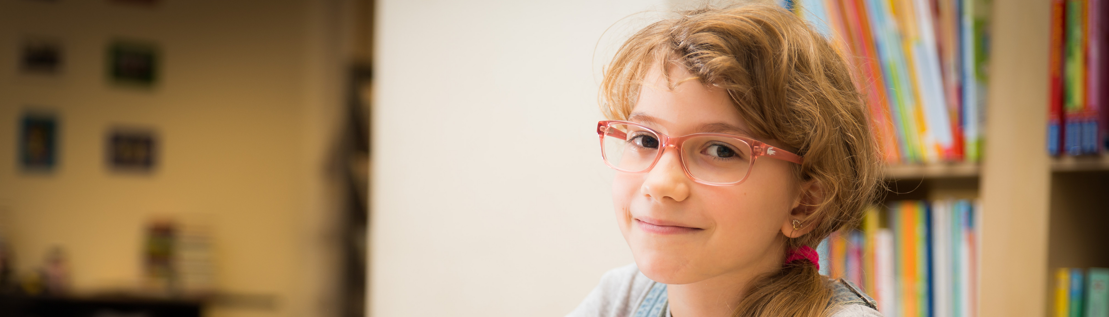 Mädchen mit roter Brille und Zopf, helle Haare, sitzt an Schreibtisch, davor Hefte, hält Stift in Hand und blickt seitlich in Kamera, im Hintergrund Bücherregal mit Büchern 