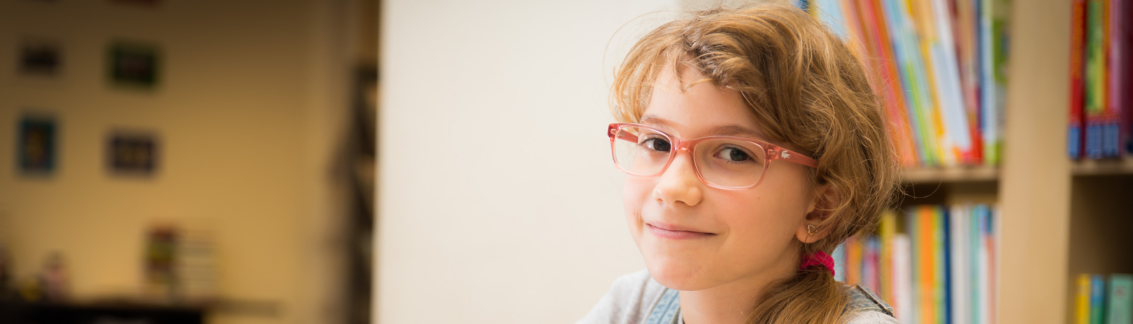 Mädchen mit roter Brille und Zopf, helle Haare, sitzt an Schreibtisch, davor Hefte, hält Stift in Hand und blickt seitlich in Kamera, im Hintergrund Bücherregal mit Büchern 