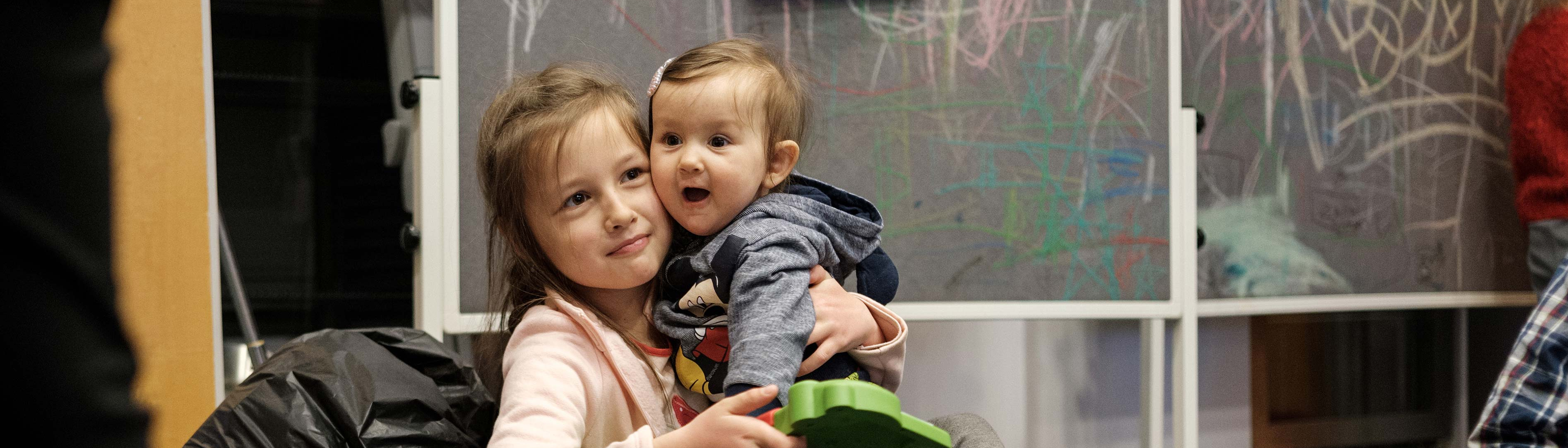 Zwei Schwestern (Kind und Baby) sitzen in Flüchtlingseinrichtung am Boden und spielen