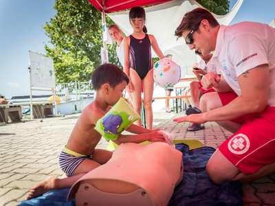 Mann mit weißem Shirt auf dem am Ärmel Wasserretung aufgedruckt ist, von der Seite fotografiert, vor ihm liegt eine Reanimationspuppe, davor hockt einer kleiner Junge in Badehose und mit Schwimmflügerl, dahinter steht Mädchen im Badeanzug und schaut zu