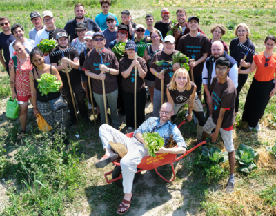 Eine Gruppe von Menschen steht auf dem Feld. Ein Mann liegt grinsend in einer Schubkarre. 