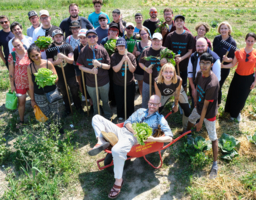 Eine Gruppe von Menschen steht auf dem Feld. Ein Mann liegt grinsend in einer Schubkarre. 