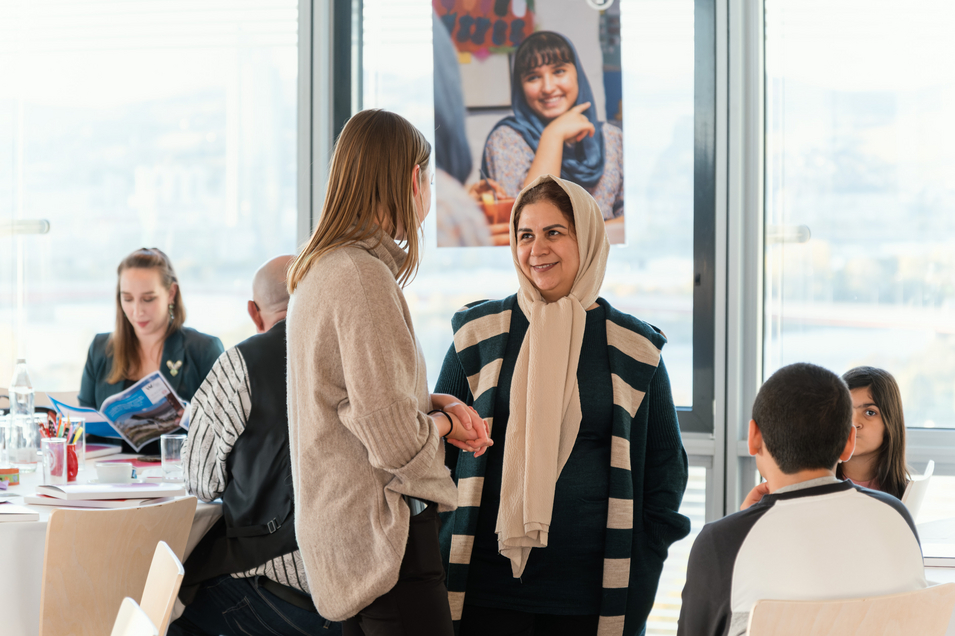 Auf dem Bild sind zwei Frauen zu sehen, die miteinander sprechen. Im Hintergrund sind Personen zu sehen, die an runden Tischen setzen.