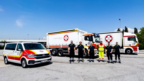 Auf dem Bild ist eine Gruppe von Personen zu sehen, die vor zwei LKWs des Samariterbund Wien stehen. Der Mann in der Mitte trägt eine Sanitäter Uniform. Im Hintergrund ist der blaue Himmel und ein paar Bäume sowie eine Brücke zu sehen.