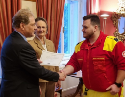 Ein Mann übergibt einem Rettungssanitäter einen Lebensretterpreis und schüttelt ihm dabei die Hand. Daneben steht eine Frau und lächelt. 