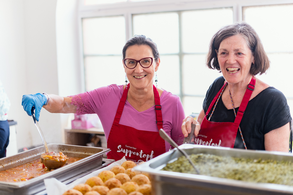 Zwei Frauen in Schürzen, die Essen in einer Küche zubereiten und lächeln.