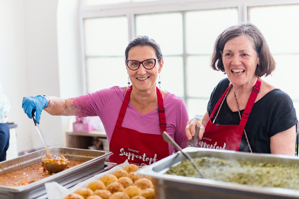 Zwei Frauen in Schürzen, die Essen in einer Küche zubereiten und lächeln.