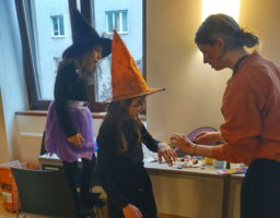 Zwei Mädchen sind als Hexen verkleidet und tragen spitze Hexenhüte. Rechts von ihnen steht eine Frau, welche die Kinder passend schminkt. Im Hintergrund ist ein Tisch mit Kinderschminke zu sehen.