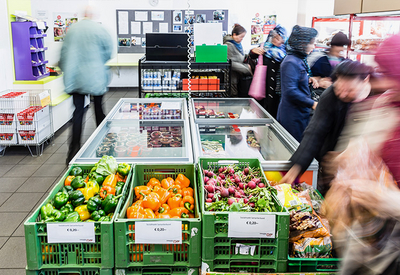 Aufnahme Sozialmarkt, Fokus auf grüne Kisten mit verschiedenem Gemüse, dahinter Tiefkühltruhe mit Glasdeckel, mehrere Personen rechts im Bild, sind alle verschwommen erkennbar, links eine Person von hinten verschwommen erkennbar, Einkaufswägen