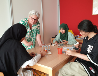 Gruppe von Mädchen sitzen am Tisch und bauen geometrische Körper, eine Frau steht daneben und erklärt.