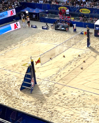 Aufnahme auf Beachvolleyball Court, keine Spieler, Sandboden, Netz, im Hintergrund Tribüne mit Werbebannern ersichtlich