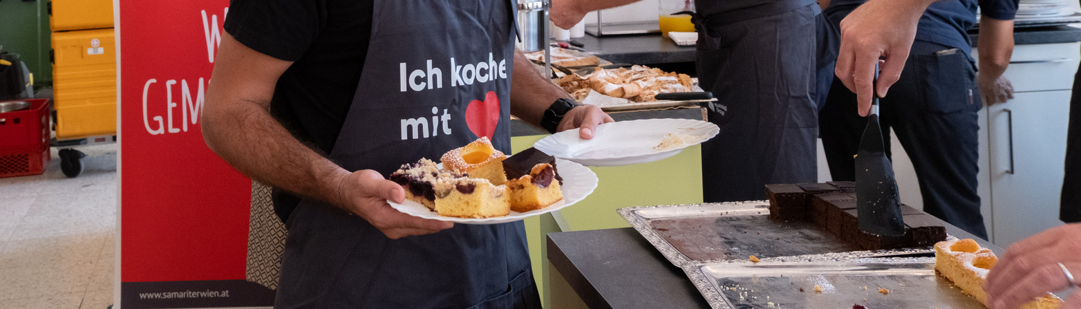 Ein lächelnder Mann in einem grauen T-Shirt und einer Schürze hält zwei Teller mit Kuchen. Im Hintergrund sind mehrere Personen beschäftigt, Essen zuzubereiten. Auf einem Banner steht „Ich koche mit...“. Der Raum ist hell und großzügig eingerichtet.