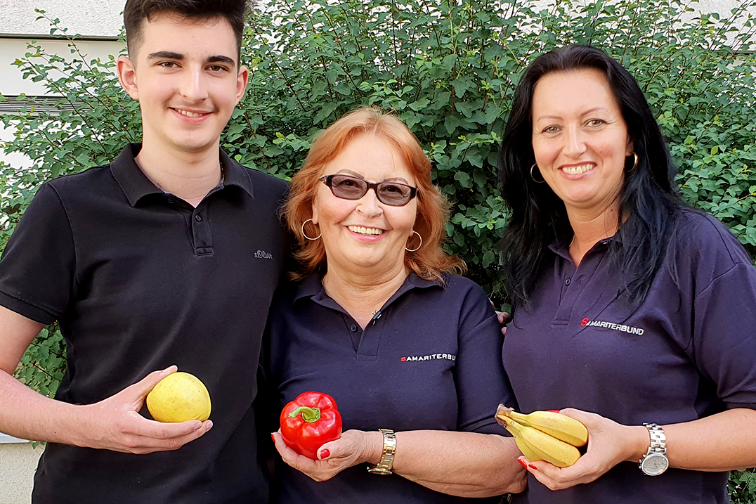 Ein Mitarbeiter und zwei Mitarbeiterinnen aus dem Sozialmarkt posieren mit jeweils einem Stück Obst in der Hand