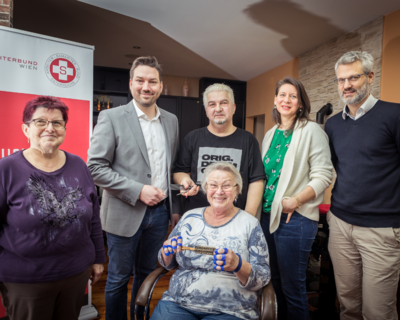 Gruppenfoto Klient:innen Sozialmarkt, Bezirksvorsteher und Friseurin, sowie Oliver Löhlein bei Gratis-Haarschnitt Aktion, im Hintergrund Samariterbund Roll-up