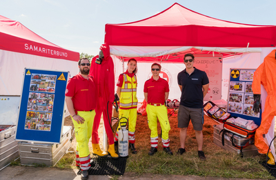 Gruppenbild, vier Personen, davon 3 in Uniformen und eine Person mit kurzer Hose und blauem Polo-Shirt vor einem Samariterbund Zelt