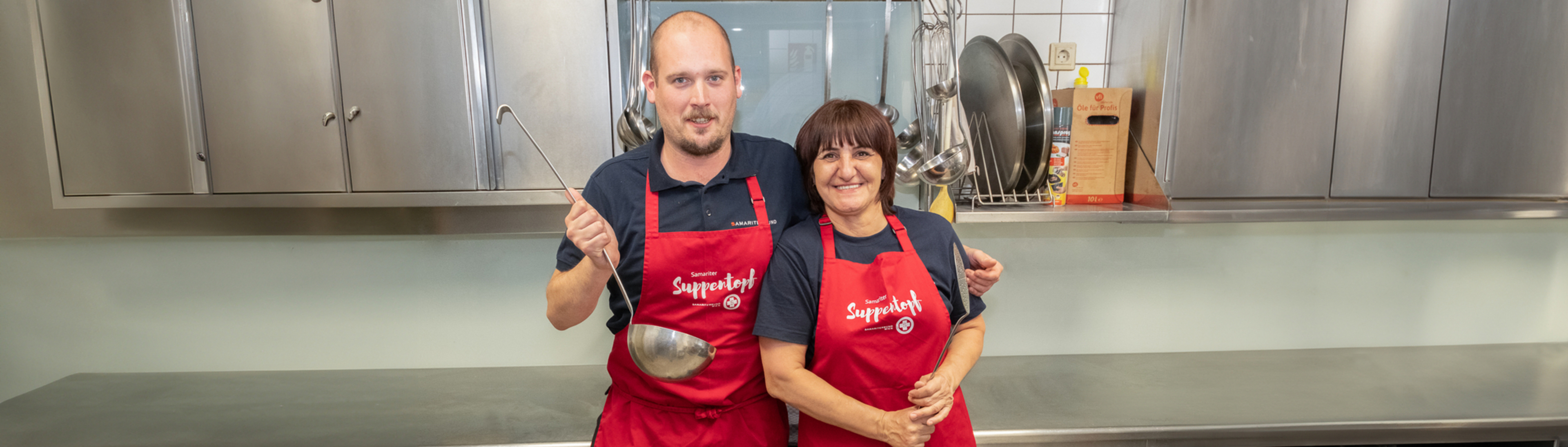 Ein Mann und eine Frau stehen nebeneinander in der Küche und lächeln. Der Mann hat einen riesigen Schöpfer in der Hand.