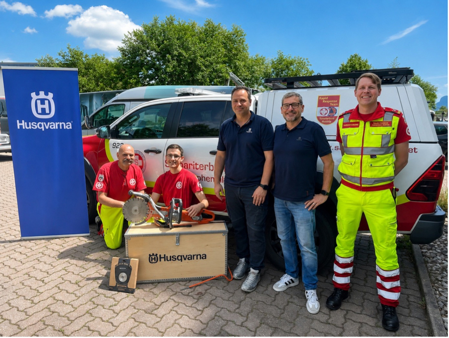 Fünf Personen stehen in einem Außengelände neben einem Fahrzeug. Zwei Personen tragen rote Shirts, während die anderen in Zivilkleidung bzw. Arbeitskleidung gekleidet sind. Vor ihnen befindet sich eine Kiste mit Werkzeugen und eine Fahne mit dem Husqvarna-Logo im Hintergrund.