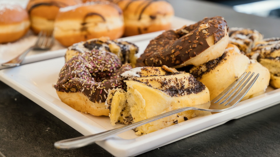 Ein Teller mit verschiedenen Donuts. Einige sind mit Schokolade und Streuseln überzogen, während andere mit einer süßen Füllung und einer Zuckerglasur versehen sind. Ein paar Gabeln liegen neben den Donuts. Im Hintergrund sind weitere Donuts sichtbar.