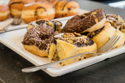 Ein Teller mit verschiedenen Donuts. Einige sind mit Schokolade und Streuseln überzogen, während andere mit einer süßen Füllung und einer Zuckerglasur versehen sind. Ein paar Gabeln liegen neben den Donuts. Im Hintergrund sind weitere Donuts sichtbar.