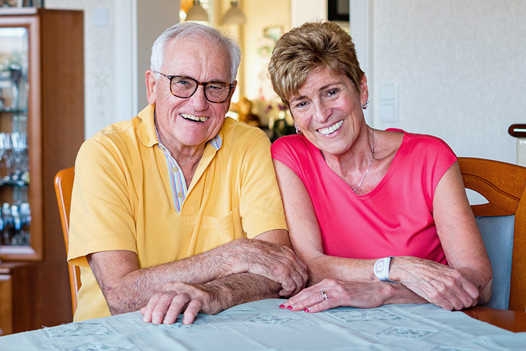 Älteres Ehepaar sitzt an Wohnzimmertisch und die Frau trägt ein Heimnotruf-Armband