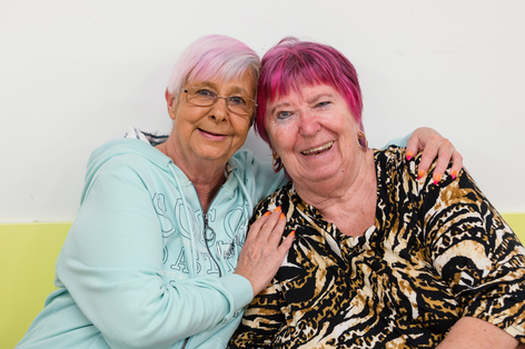 Zwei Damen mit rosa Haaren umarmen sich von der Seite und lachen in Kamera
