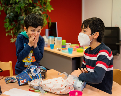 zwei Buben aus LernLEO freuen sich über Geschenke, einer hat Hände auf Mund vor lauter Freude, anderer trägt FFP2-Maske und blickt zu anderem Jungen