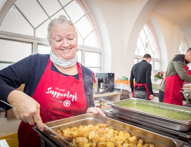 Eine ältere Dame mit roter Kochschürze bereitet Essen vor, Suppentopf, armutsbetroffene Menschen