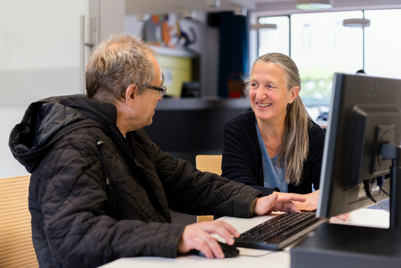 Internetcafé ZwischenSchritt – Mitarbeiterin unterhält sich mit Klient, der vor dem Computer sitzt