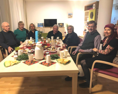 Gruppenfoto, Senior:innen von Senioren WG rund um Tisch, alle blicken in Kamera und lächeln, rundherum Wohnraumatmosphäre