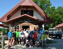 Eine Gruppe von Kindern und Betreuer:innen steht vor einer Hütte. Im Hintergrund sind Bäume und ein klarer Himmel zu sehen.