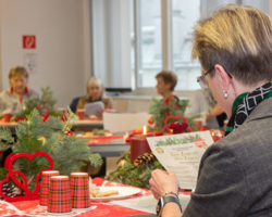 Aufnahme von Adventsjause, Person von hinten fotografiert, liest bzw. singt von einem Zettel ab, daneben Weihnachtsdeko, im Hintergrund verschwommen weitere Senioren sichtbar