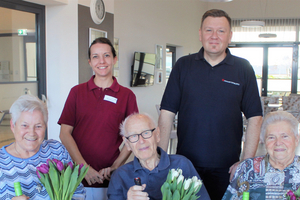 Vier Senioren halten Blumen und Geschenke, während zwei Betreuer lächeln.