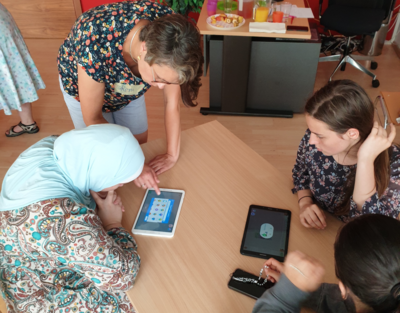 Eine Gruppe von Mädchen sitzt an einem Tisch und bedienen ein Tablet, eine Frau steht daneben und erklärt.