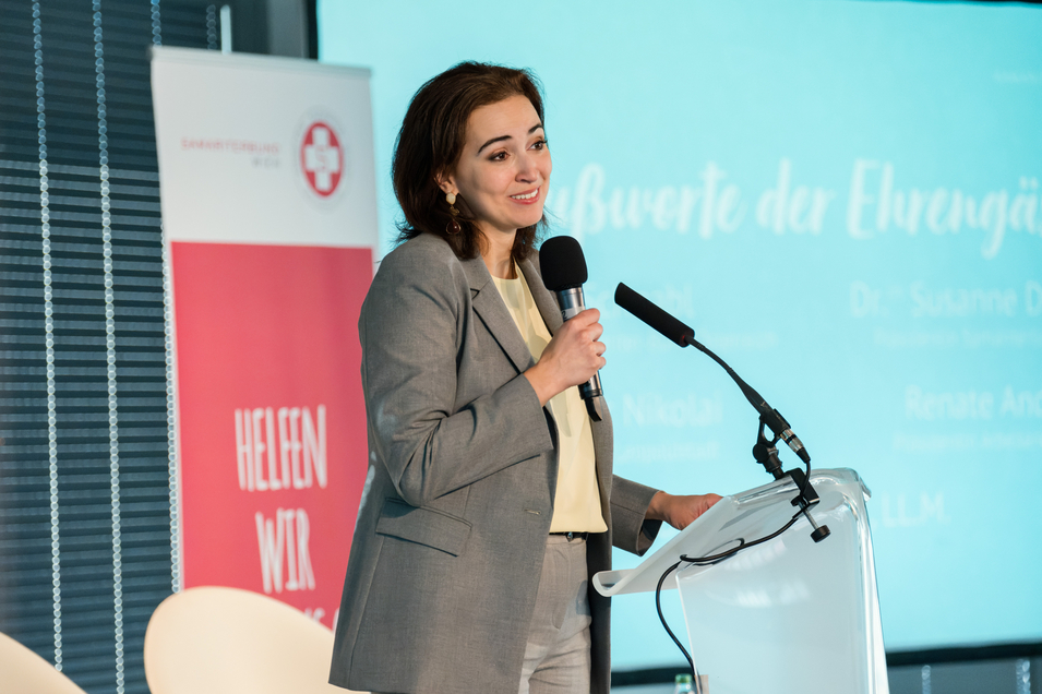 Auf dem Bild ist Alma Zadić zu sehen. Sie steht auf einer Bühne und hält ein Mikrofon in der Hand. Hinter ihr ist eine Präsentation projiziert. 