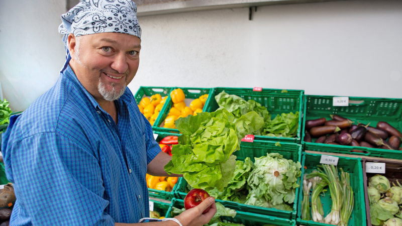 Ein Mann hält frisches Gemüse in einem Marktstand mit vielen Gemüsekisten.