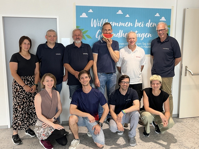 Gruppenfoto Team QSU vor Banner mit Schriftzug Willkommen bei den Gesundheitstagen, insgesamt sind 10 Personen zu sehen, 6 Personen stehen in der hinteren Reihe, vier Personen knien davor, alle lächeln in Kamera, eine Person hält eine Wassermelone vor dem Gesicht 