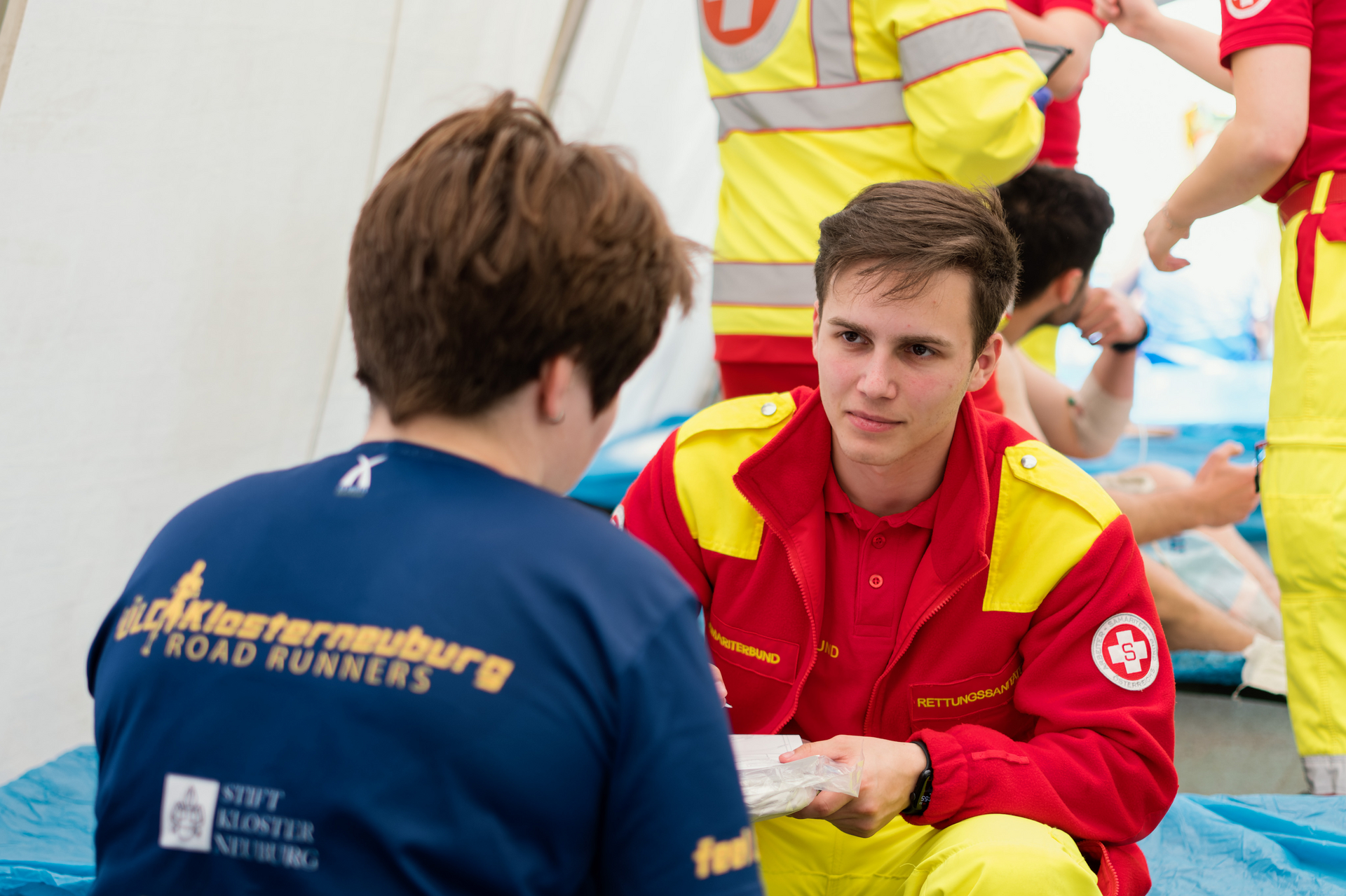 Ein Rettungssanitäter des Samariterbundes versorgt in einem medizinischen Zelt eine Teilnehmerin des Marathons, die ein Shirt der 'Klosterneuburg Road Runners' trägt. Im Hintergrund sind weitere Rettungskräfte und Patienten zu sehen, die versorgt werden.