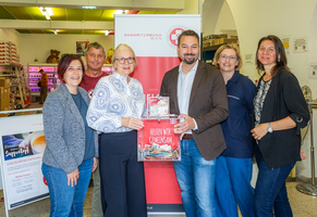 Gruppenfoto, 6 Personen vor bzw. neben Roll up vom Samariterbund Wien mit Schriftzug Helfen wir gemeinsam, Personen in der Mitte halten Spendenbox mit Geld, alle blicken in Kamera und lächeln, im Hintergrund Sozialmarkt