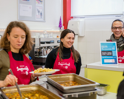 Mitarbeiter:innen Firma Insight bei Samariter Suppentopf, beim Ausgeben von Essen, eine Person hält Schöpflöffel in der Hand und gibt auf Teller, zwei weitere Personen im Hintergrund