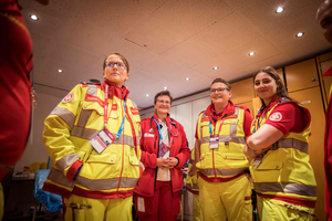Fünf Personen in Rettungsdienstkleidung stehen in einem Raum. Die Frauen tragen rote und gelbe Uniformen mit Logos. Sie blicken lächelnd in die Kamera und scheinen ein Team zu bilden, während sie sich im inneren eines Gebäudes befinden.