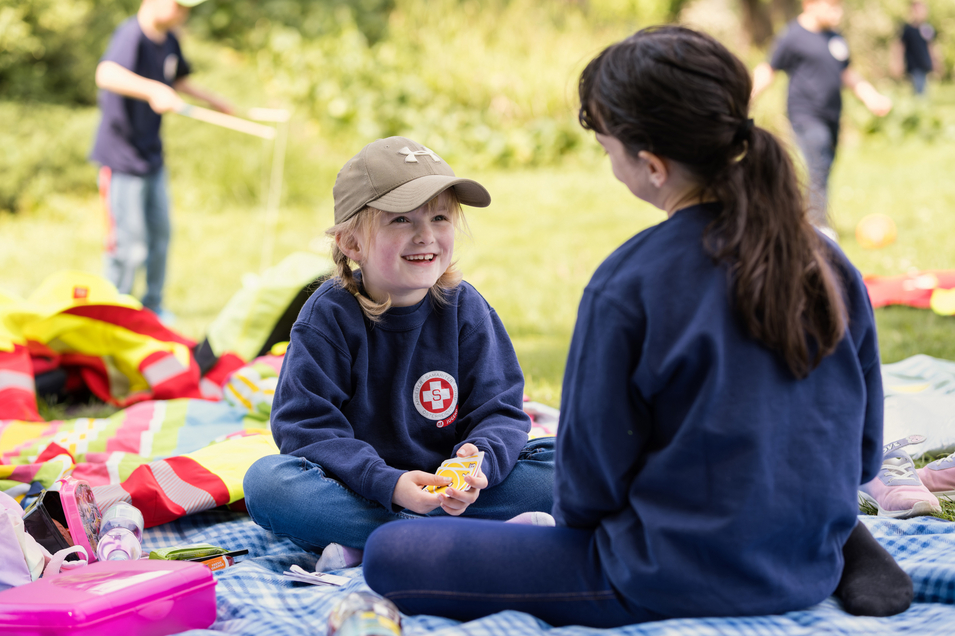Zwei Kinder der Samariterjungend spielen Karten auf einer Picknickdecke im Park und lachen sich dabei an