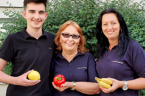 Ein Mitarbeiter und zwei Mitarbeiterinnen aus dem Sozialmarkt posieren mit jeweils einem Stück Obst in der Hand