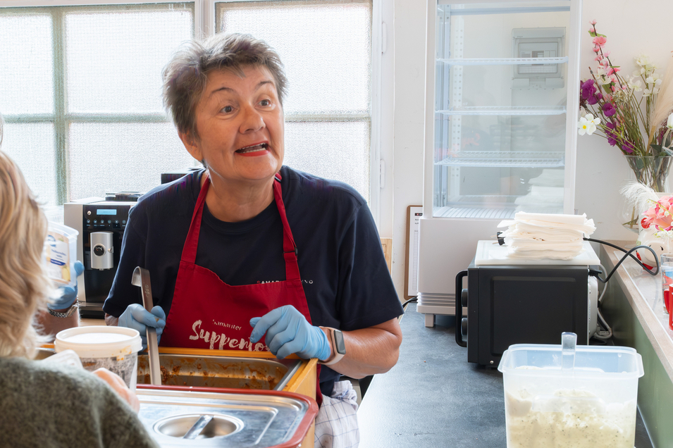 Eine Frau mit Schürze spricht und serviert Essen in einer Küche.