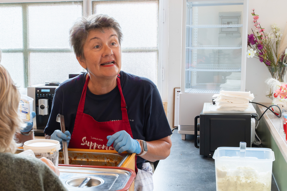 Eine Frau mit Schürze spricht und serviert Essen in einer Küche.