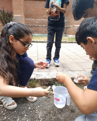 Vier Kinder, drei von ihnen sitzen, ein Kind steht. Sie sind im Freien und führen ein Experiment mit einem Messbecher durch.