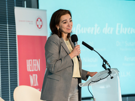 Auf dem Bild ist Alma Zadić zu sehen. Sie steht auf einer Bühne und hält ein Mikrofon in der Hand. Hinter ihr ist eine Präsentation projiziert. 