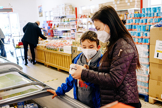 Mutter und Sohn stehen vor Kühltruhe im Sozialmarkt und Mutter hält ein Produkt in der Hand