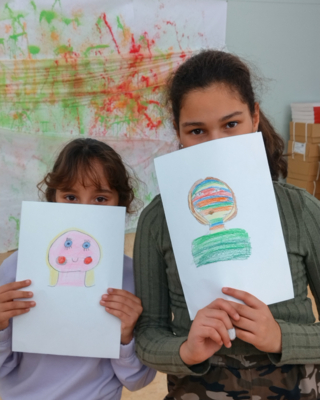 zwei unterschiedlich große Mädchen, stehen nebeneinander und halten ein A4 großes Blatt mit einer Zeichnug vor ihren Gesichtern